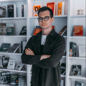 Studentas, trumpais tamsiais plaukais, tamsiais akiniais, dėvintis tamsius marškinius, sukryžiavęs rankas, stovintis knygų lentynų fone