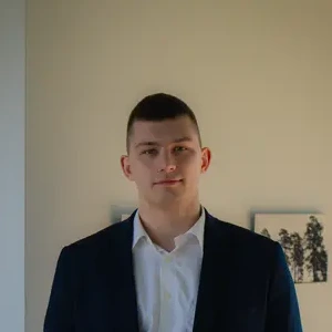 Laimingai atrodantis vyras trumpais plaukais, vilkintis baltus mar&scaron;kinius, mėlyną &scaron;varką, &scaron;viesus fonas su kabančiais paveikslais.